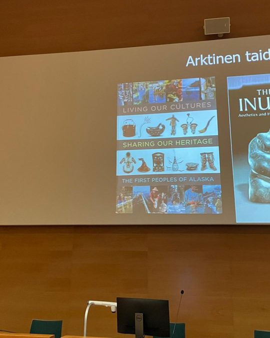 Timo Jokela talar i en auditorium, power point: en affisch eller bok med text Living our cultures - sharing our heritage - the first peoples of Alaska och bok: The way of Inuit art.